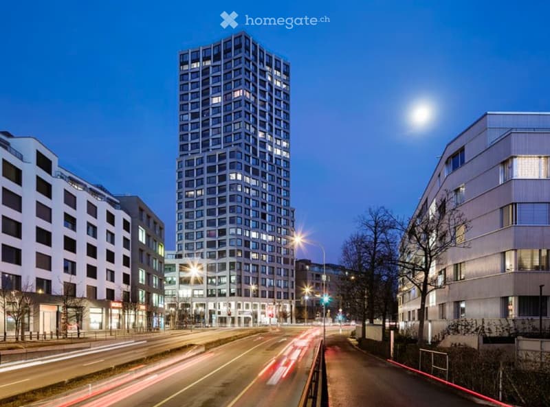 Modern high-rise building with illuminated windows, surrounded by other contemporary structures and a busy street with light trails from moving vehicles.