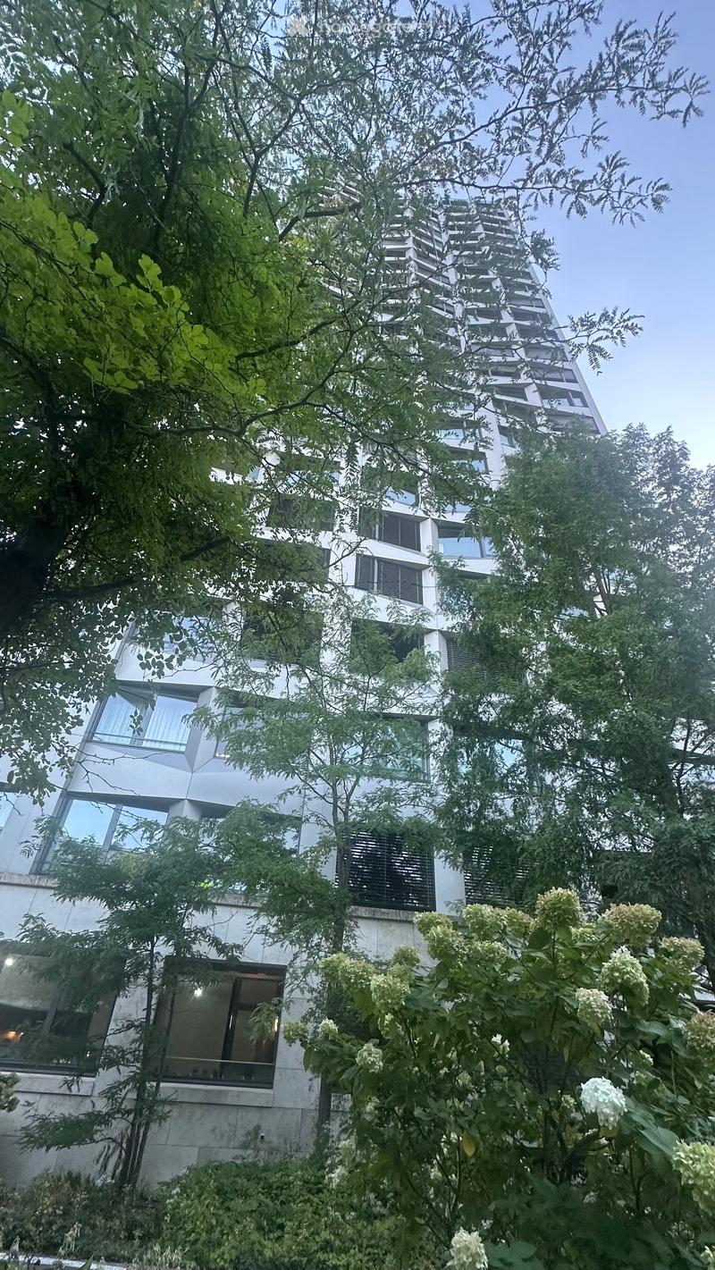 Modern high-rise building with large windows and balconies, surrounded by lush greenery and trees.