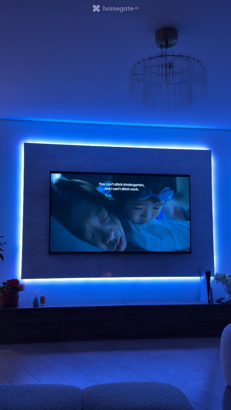 Modern living room with a large flat-screen TV mounted on a wall, illuminated by ambient blue lighting, a dark wooden TV stand, and decorative items including a flower vase and small ornaments.
