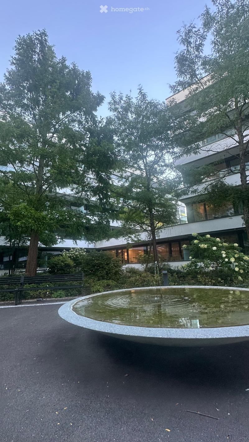 A modern building with a fountain in the foreground, surrounded by trees and greenery.