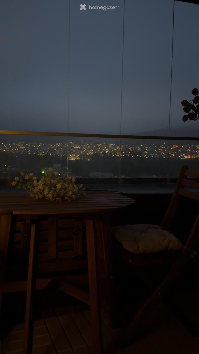 Balcony with a wooden table, chairs, and a flower arrangement, overlooking a cityscape at night.