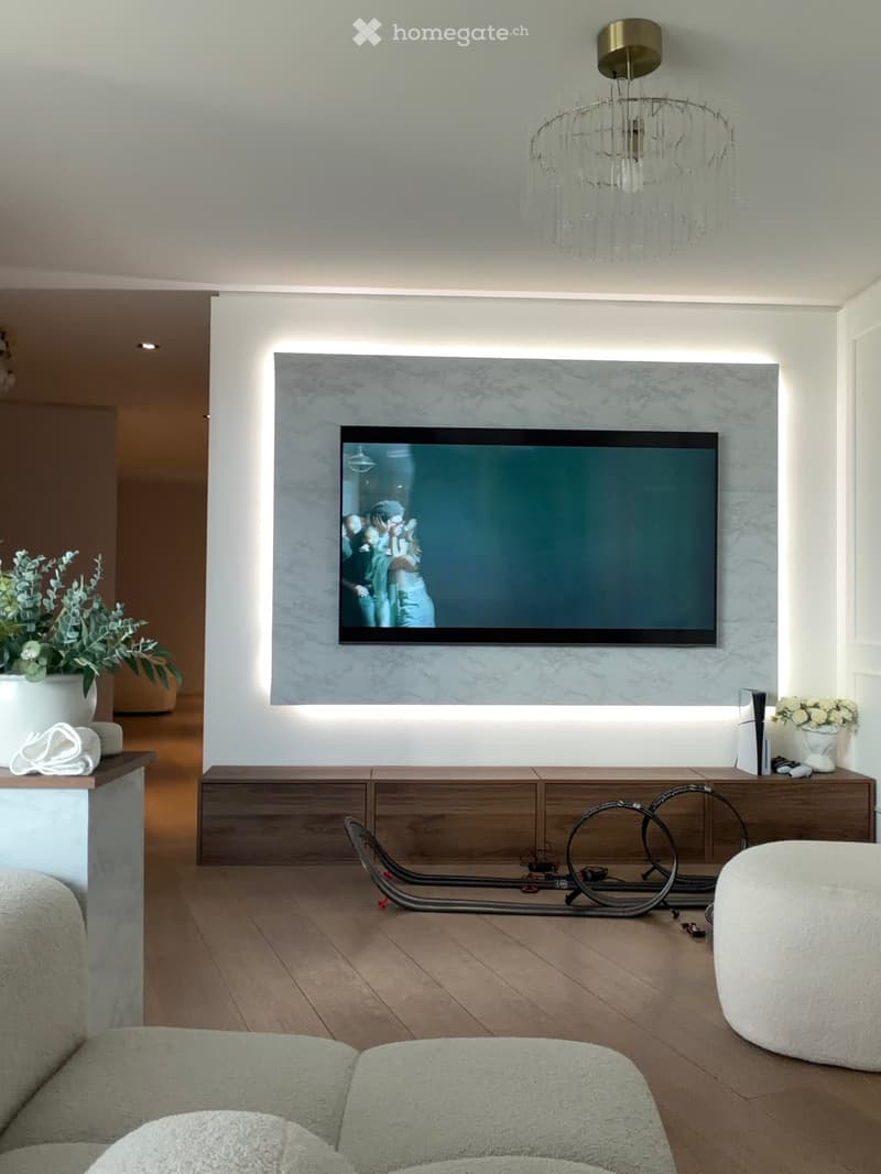 Modern living room with a large flat-screen TV mounted on a marble wall, a sleek wooden entertainment unit, a plush sofa, ottoman, and decorative vases.