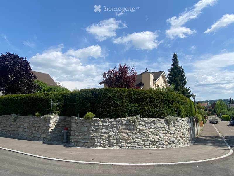 A two-story house with a stone wall and hedge in front, surrounded by trees and a clear blue sky.