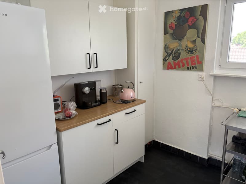 Modern kitchen with white cabinets, wooden countertop, and various appliances including a coffee maker, kettle, and refrigerator. A poster is on the wall, and a cart is near the window.