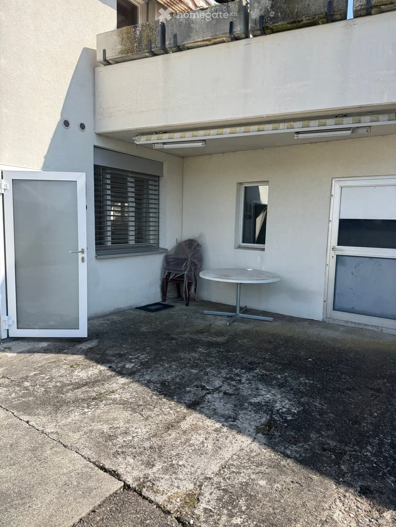 Exterior of a building with a white wall, two doors, a window with blinds, a table, and a chair covered with plastic.