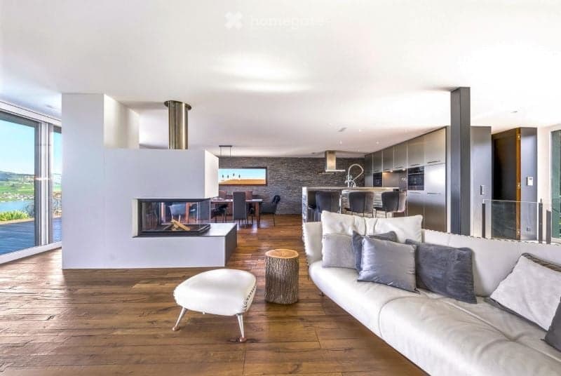 Open-plan living and dining area with a modern fireplace, large windows offering a scenic view, a white sofa with grey cushions, an ottoman, a dining table with chairs, and a kitchen island with bar stools.