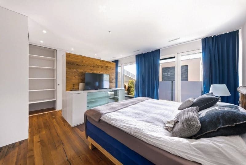 Modern bedroom with wooden flooring, large window with blue curtains, a neatly made bed with pillows and blankets, a nightstand with a lamp, a television on a cabinet, and built-in shelving unit.