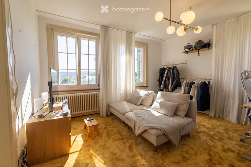 Bright living room with large windows, a grey sofa, a wooden side table, a television, and a clothing rack with hanging clothes.