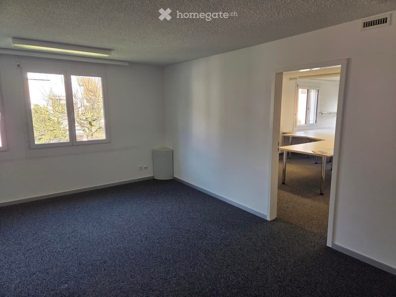 Empty room with grey carpet, white walls, and large windows. Adjacent room contains a table and chairs.