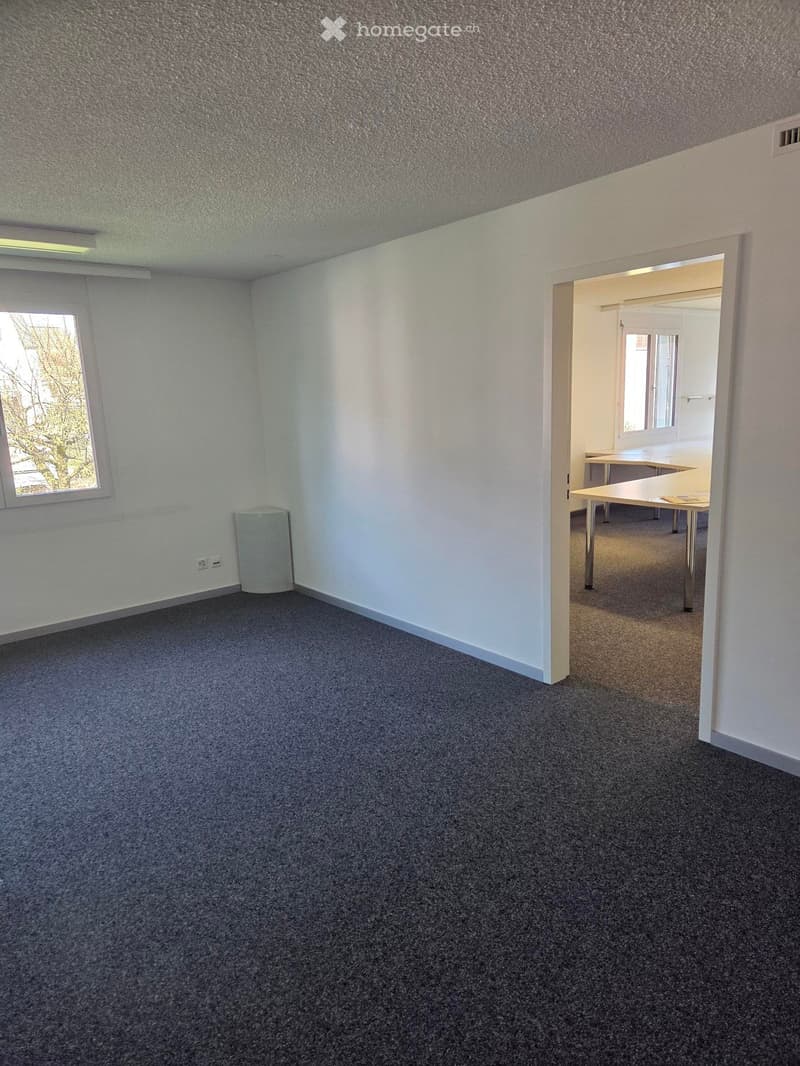 Empty room with grey carpet, white walls, and a window. Adjacent room contains a table and chairs.