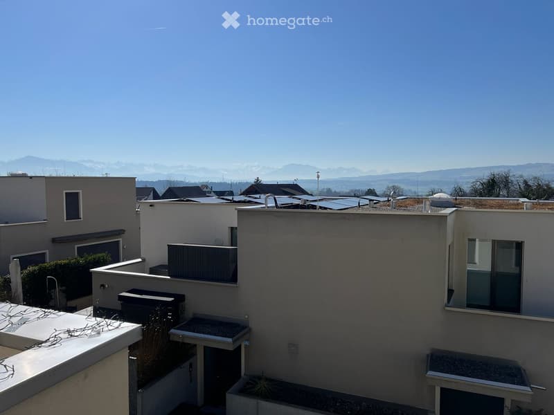 Building. Modern multi-story residential building with flat roofs, solar panels, and surrounding greenery, overlooking a scenic mountain landscape.