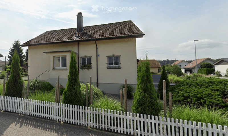 House. Single-story house with beige exterior, tiled roof, chimney, and white picket fence surrounding a garden with tall conifers and shrubs.