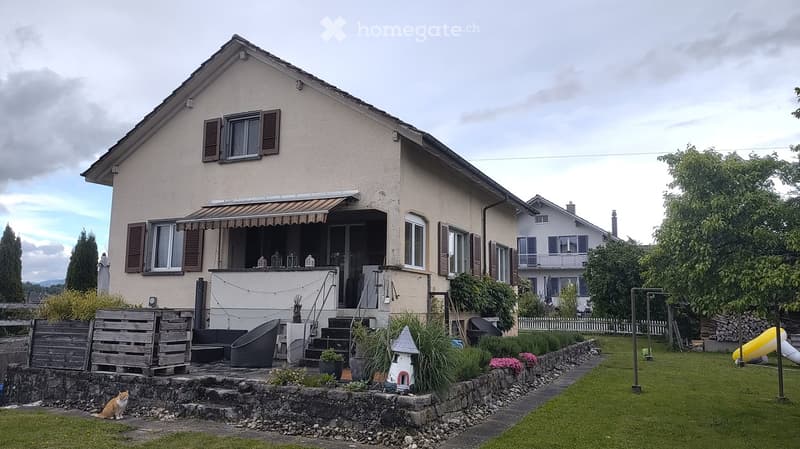House. Two-story house with beige facade, multiple windows, covered balcony, and well-maintained garden with seating area, plants, and a cat.