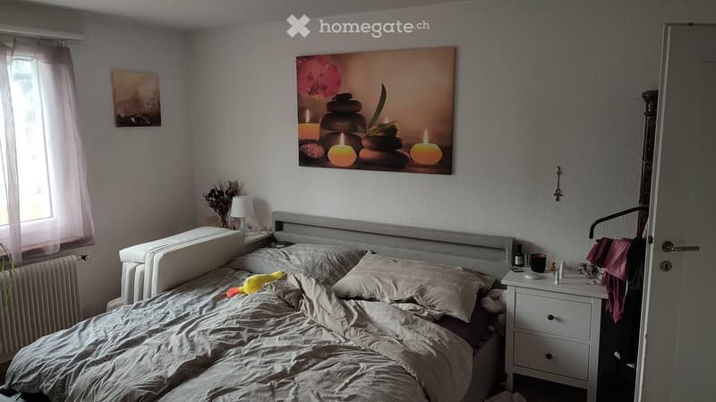 Bedroom. Cozy bedroom with a large bed, nightstand, lamp, and a large picture frame above the bed. The room features a window with a curtain, a radiator, and a small plant on the nightstand.