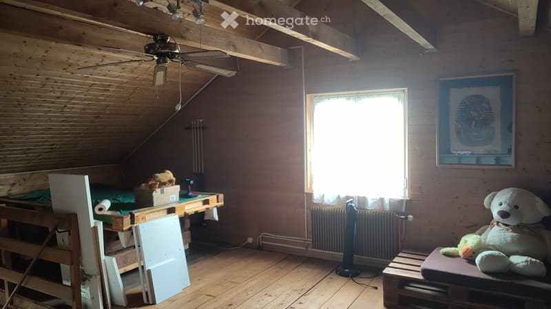 Furnished Room. The room has wooden walls and ceiling, a large window with curtains, a table with a laptop, a radiator, a fan, a picture frame, and a large stuffed animal on a stool.