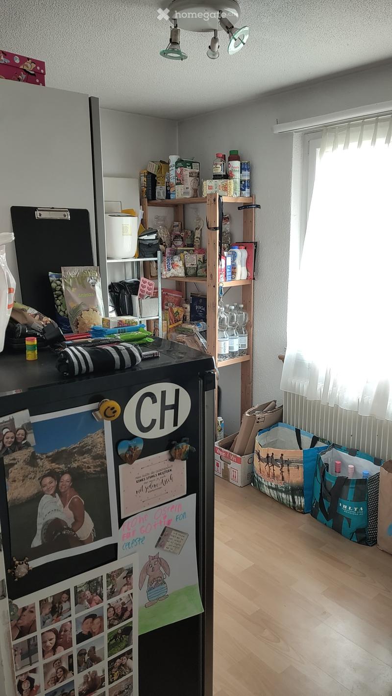 Furnished Room. A small, furnished room with a refrigerator, shelves filled with various items, and a wooden floor. The refrigerator is decorated with photos and notes.