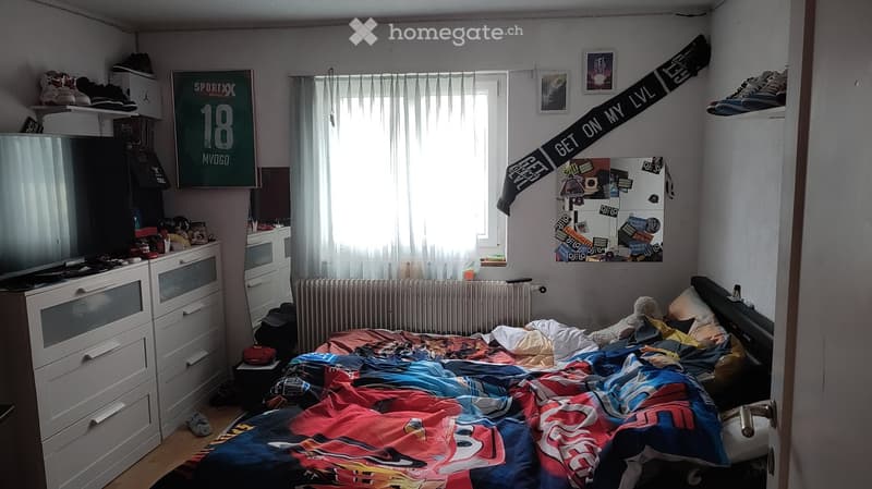 Bedroom. A cozy bedroom with a bed covered in a colorful blanket, a nightstand with a mirror, shelves with shoes and items, a radiator, a window with curtains, and posters on the walls.