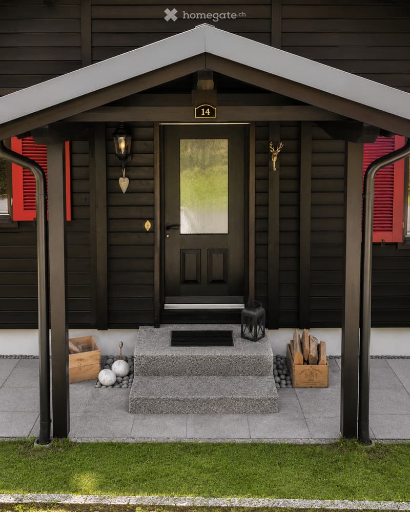Front entrance of a house with a covered porch, black door, lantern-style light fixture, decorative deer head, and stacked firewood crates.