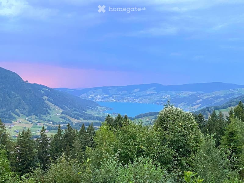 Panoramic view of a valley with a lake, surrounded by forested hills and mountains under a partly cloudy sky.