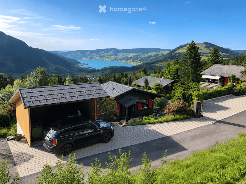 A modern two-story house with a black roof and red shutters, featuring a garage with a parked car, surrounded by lush greenery and a scenic mountain view.