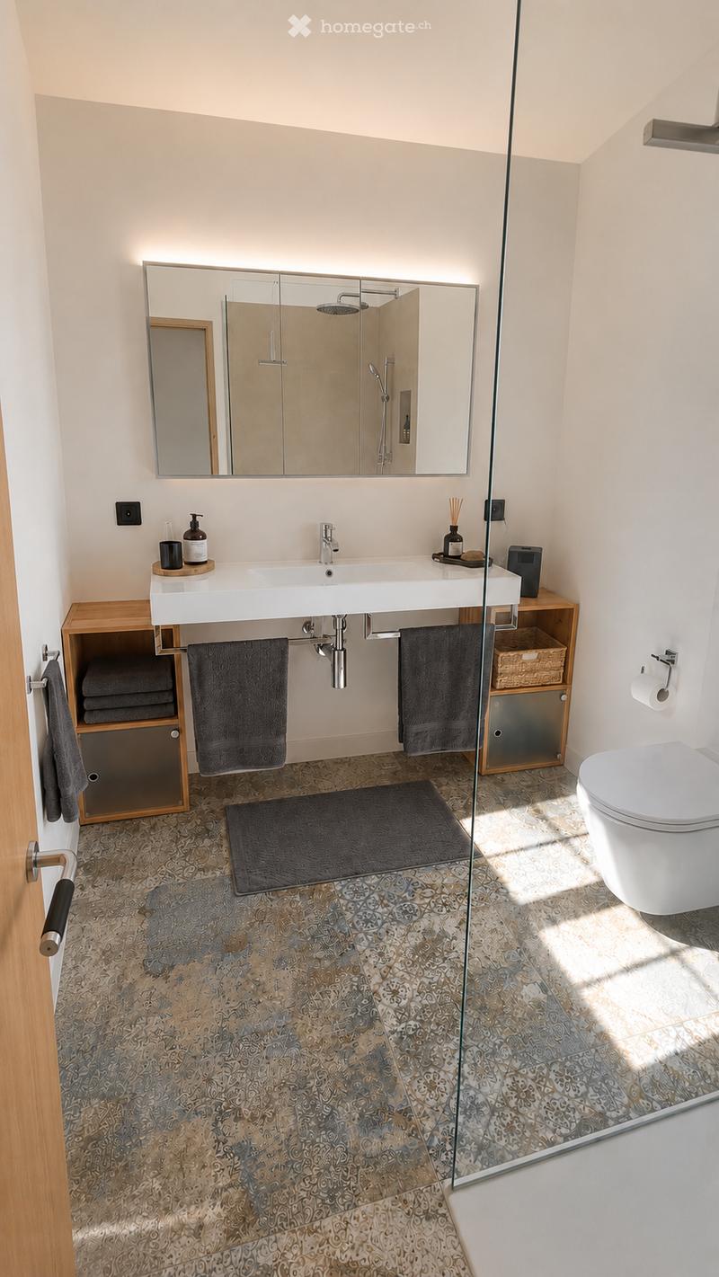 Modern bathroom with a large mirror, white sink, dark towels, and a glass-enclosed shower. The floor features a patterned tile design, and the space includes a toilet and a small wooden cabinet.