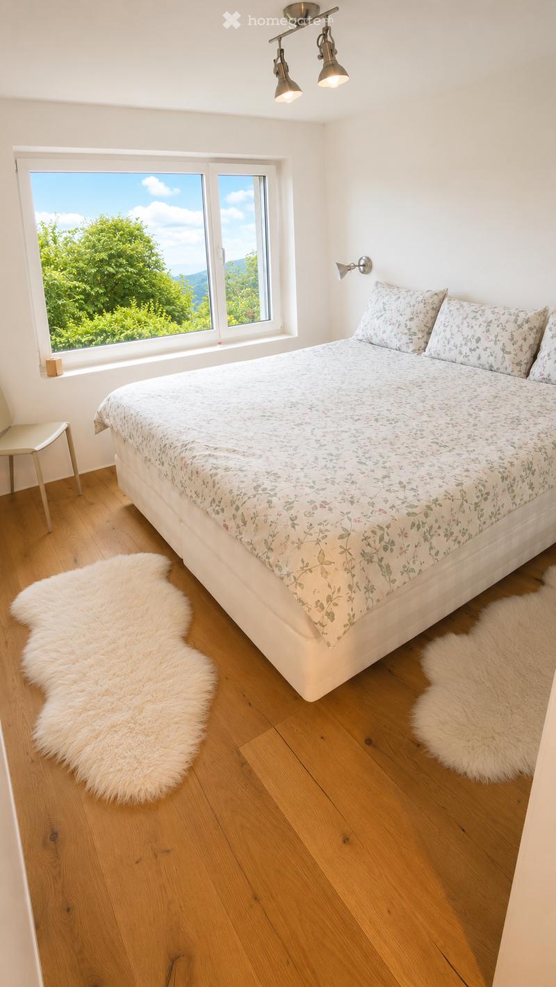 Bright bedroom with wooden flooring, large window offering a scenic view, a neatly made bed with floral bedding, a white fur rug, and a small chair near the window.