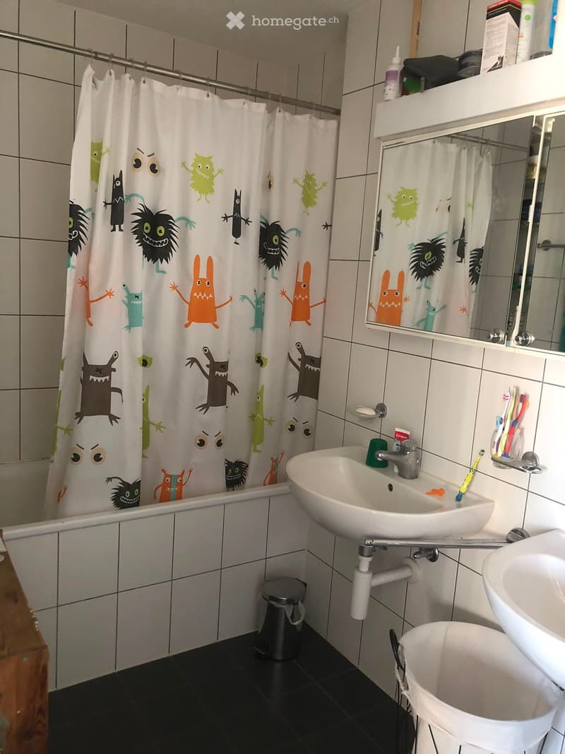 Bathroom with a white sink, mirror, bathtub with a colorful monster-themed shower curtain, and a toothbrush holder with toothbrushes.