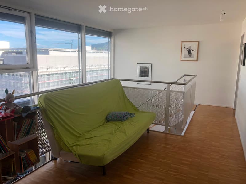 A modern living room with a green futon sofa, a small bookshelf, a toy, and picture frames on the wall. Large windows offer a scenic view.