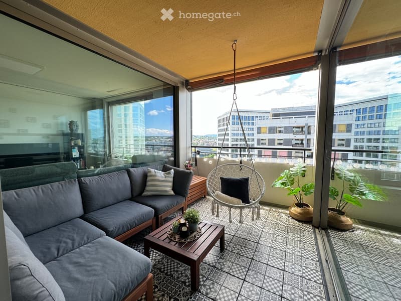 Balcony with grey sectional sofa, wooden coffee table, wicker hanging chair, and potted plants, overlooking cityscape.