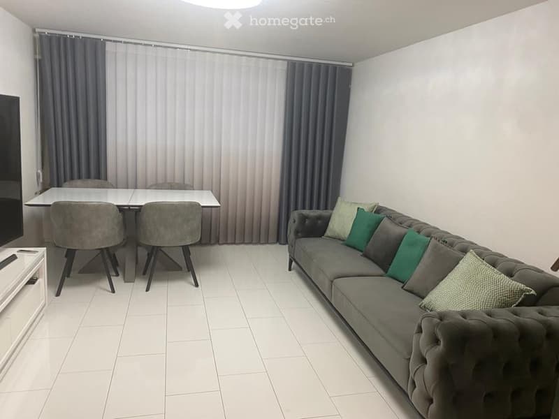 Modern living room with a grey sofa, multiple pillows, a white dining table with four chairs, and a television on a white stand.