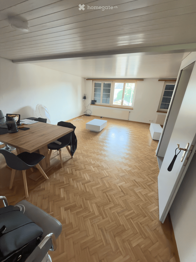 Bright room with herringbone wooden flooring, large windows, and modern furniture including a wooden table, chairs, and small white stools.