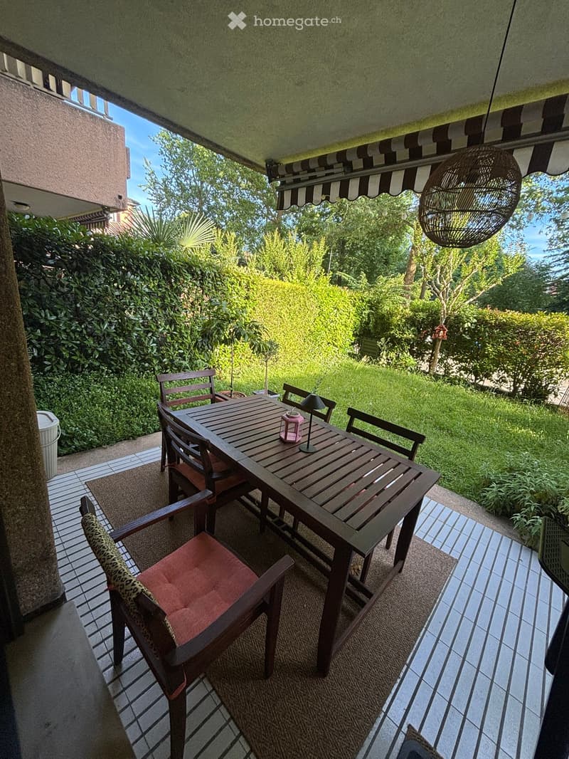 Balcony with wooden table, chairs, and a hanging lamp, overlooking a green garden.