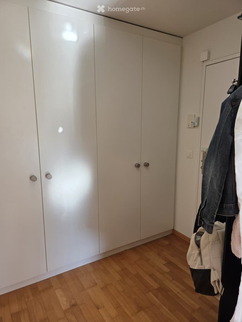 Empty room with light-colored wooden flooring and white built-in wardrobes. A coat and bag are hanging on a rack near the door.