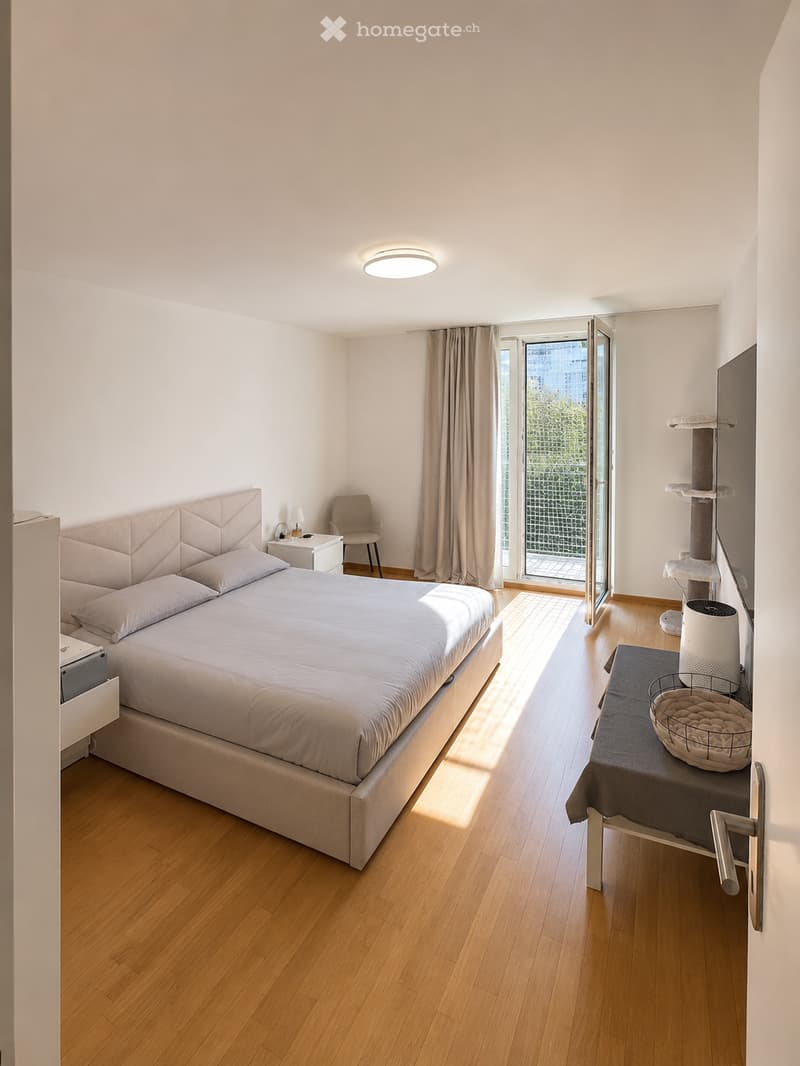 Modern bedroom with a large bed, nightstand, chair, cat tree, and a small table with a cat bed on it. The room features wooden flooring, large windows with curtains, and a sleek, contemporary design.