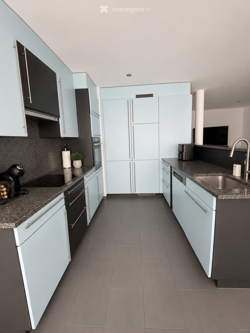 Modern kitchen with light blue cabinets, dark countertops, stainless steel appliances, and a central island.