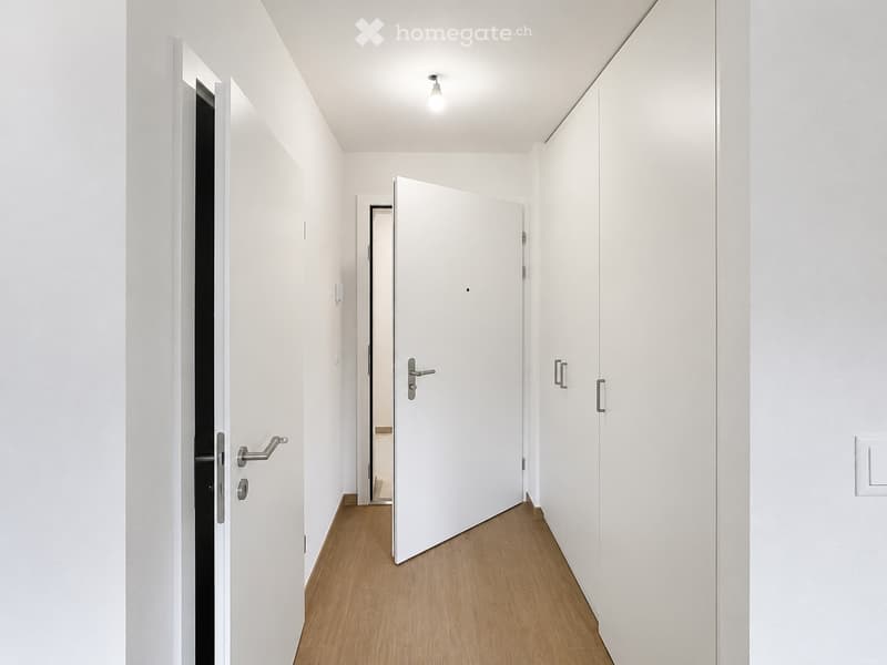 A clean, modern hallway with white walls, light wooden flooring, and several closed white doors with silver handles. The ceiling has a single recessed light fixture.