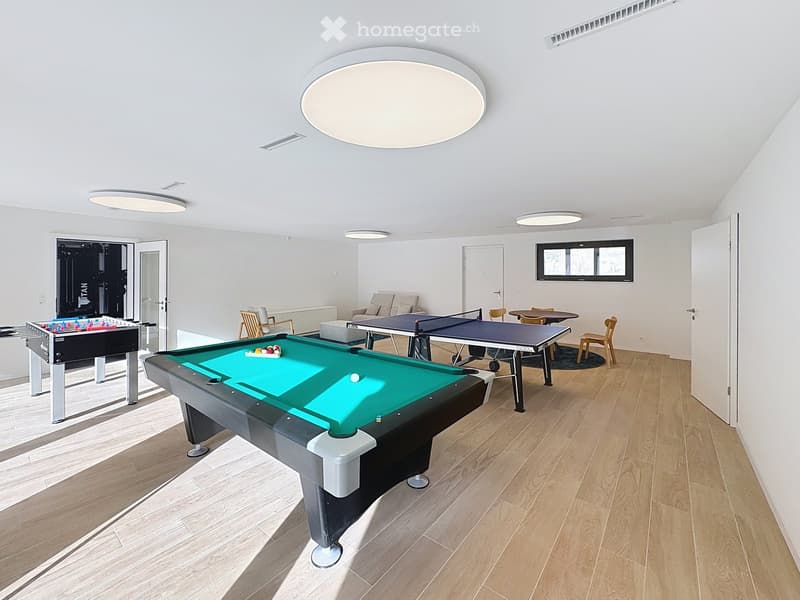 A spacious room with wooden flooring, featuring a pool table, a table tennis table, a sofa, chairs, and a screen on the wall.