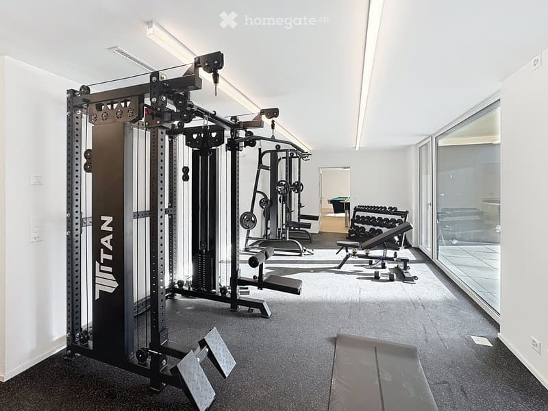 Modern gym room with weight machine, bench, dumbbells, large windows, and sleek design.