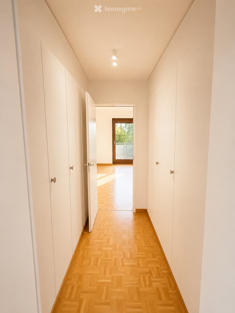 A long, narrow hallway with light-colored walls, wooden herringbone flooring, and recessed ceiling lights. The hallway features several closed white doors on both sides and leads to an open doorway at the end.