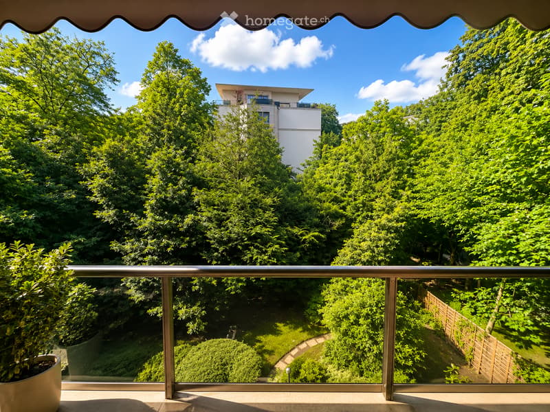 Balcony with metal railing, overlooking a lush green garden and a modern white building in the background.