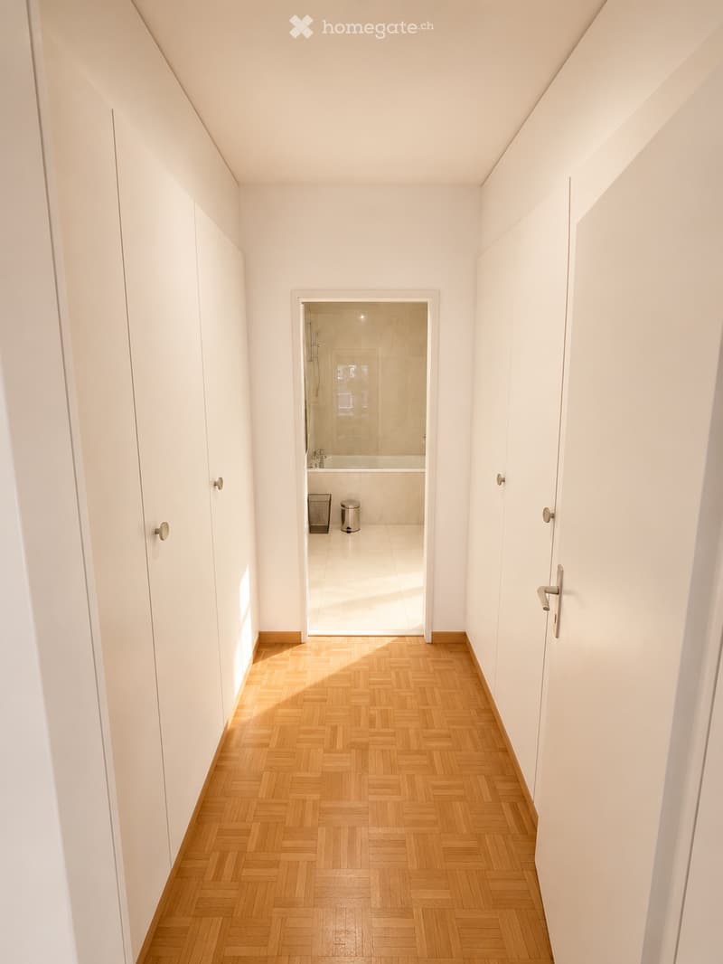 A narrow hallway with light wooden herringbone flooring and white walls, leading to a modern bathroom with a glass-enclosed shower and a bathtub.