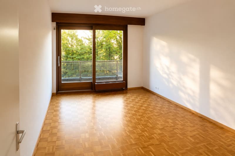 Empty room with wooden parquet flooring, white walls, and a large sliding glass door leading to a balcony with a metal railing. Sunlight streams in, casting shadows on the walls.