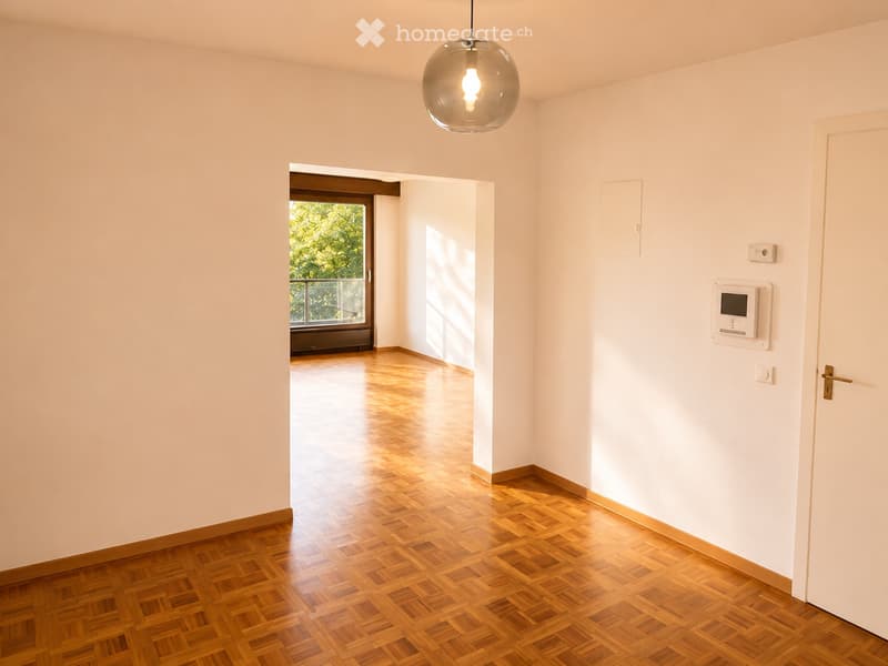 Empty room with light-colored walls, wooden parquet flooring, and a pendant light fixture. A doorway leads to a balcony area with a view of greenery.