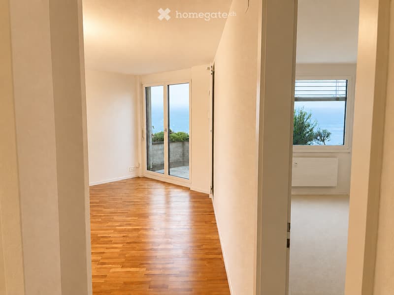 Empty hallway with wooden flooring, white walls, and large windows offering a view of greenery and water. Features a sliding glass door leading to an outdoor area.