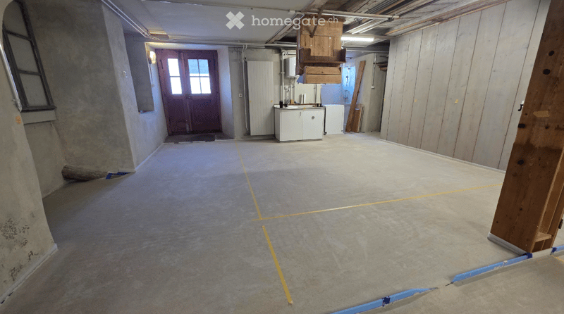 Empty basement room with concrete floors, exposed wooden beams, and a small utility area with cabinets and a sink.
