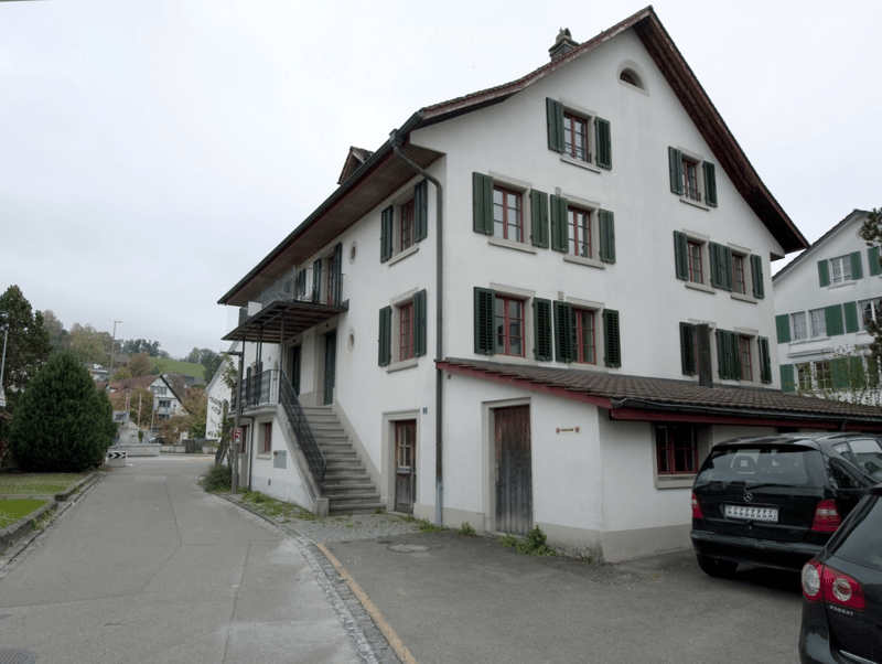 Two-story white building with green shutters and a balcony, located on a street with parked cars.
