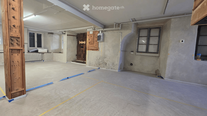 Empty room with bare concrete walls, wooden floor, and minimal fixtures. Exposed pipes and a wooden door frame are visible.