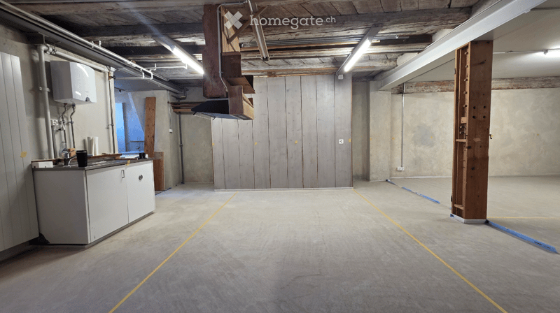Empty room with concrete walls and floor, exposed wooden beams, and a small white cabinet with a sink and faucet. No furniture or equipment.