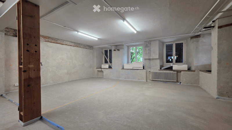 Empty room with bare walls, concrete floor, and exposed wooden beam. Windows and radiator are visible, but no furniture or equipment.