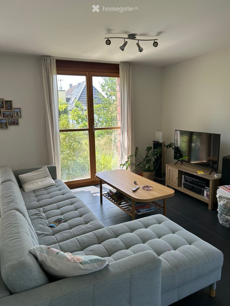 Modern living room with a grey sectional sofa, wooden coffee table, flat-screen TV on a wooden stand, and large windows with curtains. The room features a ceiling fan with spotlights and a potted plant near the TV stand.